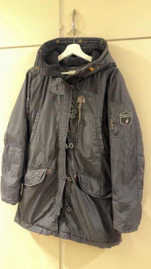 Peak performance  - Snygg mörkgrå parkas med stor huva och dragkedja samt knappar framtill. Jackan har två stora fickor med lock, bröstficka med dragkedja och patch på ärmen. Fodrad insida och justerbar nederkant. Perfekt för kalla dagar och streetwear-stil.