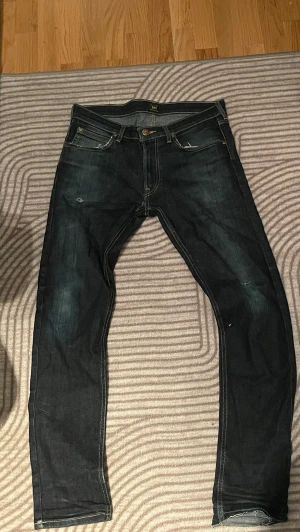Mörkblå  jeans från Lee med slitningar  - Säljer ett par mörkblå jeans från Lee med klassisk femficksmodell och guldfärgade detaljer. Jeansen har lätt slitna partier och kontrastsömmar som ger en cool look. Perfekta för dig som gillar en smal passform och tidlös stil. Snygga slitningar! Storlek 31W 32L