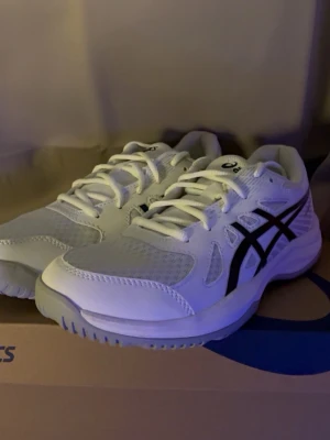 Vita Asics sneakers - Snygga vita sneakers från Asics med klassiska svarta ränder på sidorna. Skorna har en låg profil, vit sula och ovandel i mesh och syntetmaterial för bra andningsförmåga. Perfekta för dig som gillar sportig och stilren look.