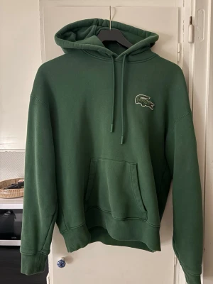 Grön hoodie från Lacoste Loose Fit - Säljer en grön hoodie från Lacoste. Hoodien har en klassisk huva med dragsko, stor magficka och den ikoniska krokodilloggan broderad på bröstet. Unisex och tillverkad i mjukt material som är skönt att bära.