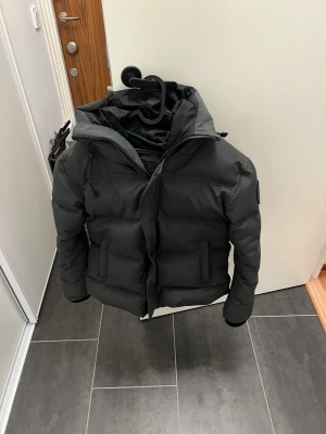 Svart dunjacka från Canada Goose - Säljer en svart dunjacka från Canada Goose med klassisk patch på ärmen. Jackan har huva, dragkedja och knappar framtill samt två fickor. Perfekt för kalla vinterdagar och riktigt snygg streetstil. Har kvar alla taggar och lappar till jackan 