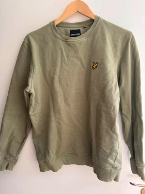 Olivgrön sweatshirt från Lyle & Scott - Olivgrön sweatshirt från Lyle & Scott med rund halsringning och klassisk gul logga på bröstet. Tröjan har ribbade muddar vid ärmslut och nederkant, samt en avslappnad passform. Perfekt för en casual och stilren look.