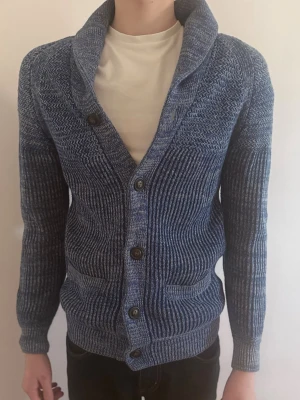 Blå stickad kofta med krage - Mysig blåmelerad stickad kofta med krage och knäppning framtill. Två framfickor och ribbade muddar vid ärmslut och nederkant. Perfekt för lager på lager och en avslappnad stil.