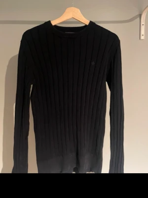 Svart ribbstickad tröja Morris - Svart ribbstickad tröja från Morris med rund halsringning och diskret logga på bröstet. Tröjan har långa ärmar och är tillverkad i ett mjukt stickat material som passar perfekt till höst och vinter. Enkel och stilren design som funkar till det mesta.