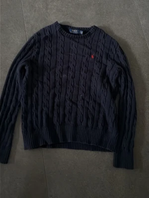 Mörkblå kabelstickad tröja Polo Ralph Lauren - Kabelstickad mörkblå tröja från Polo Ralph Lauren med rund halsringning och ribbade muddar. Tröjan har det klassiska röda broderade logot på bröstet och är tillverkad i mjukt material. Perfekt för en stilren och avslappnad look.