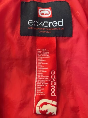 Ecko Red jeansjacka med fuskpäls - Snygg blå jeansjacka från Ecko Red med röd innerfoder och avtagbar huva med fuskpäls. Jackan har flera dragkedjeförsedda fickor och coola sömdetaljer. Perfekt för dig som vill ha en streetwear-inspirerad look. Liten lagning på insidan av jackan vid höger axel (se bild)