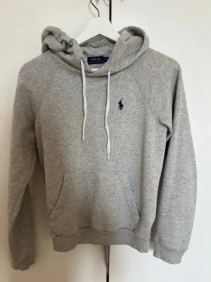 Grå hoodie från Polo Ralph Lauren - Snygg ljusgrå hoodie från Polo Ralph Lauren med klassisk marinblå logga broderad på bröstet. Hoodien har huva med vita dragsnören och en stor magficka framtill. Tunnt material och hoodien är o storlek XS