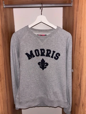 Grå sweatshirt från Morris - Snygg grå sweatshirt från Morris med svart broderad logga och dekorativ lilja på bröstet. Tröjan har rund halsringning, långa ärmar och ribbade muddar vid ärmslut och nederkant. Perfekt för en avslappnad och stilren look.