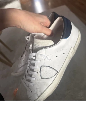 Vita Philippe Model sneakers - Snygga vita sneakers från Philippe Model med blå detalj på hälen och klassisk logga på sidan. Skorna har vita skosnören, perforerade hål vid sidan och en robust gummisula. Tillverkade i läder med mockadetaljer på plösen för en clean och stilren look.