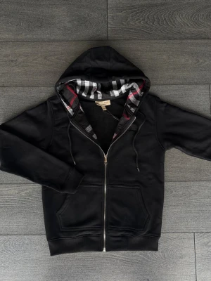 Svart hoodie från Burberry med dragkedja - Snygg svart hoodie från Burberry med dragkedja framtill och klassiskt rutigt foder i huvan i svart, vitt och rött. Hoodien har två fickor fram och justerbar dragsko i huvan. Perfekt för en stilren och avslappnad look.