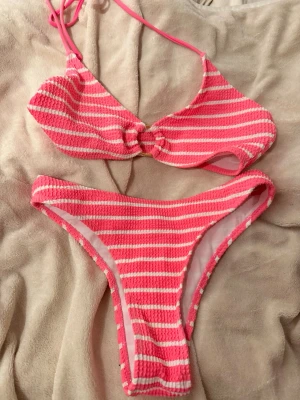 Rosa och vit randig bikini - Säljer en söt bikini i rosa och vitt med randigt mönster. Överdelen har knyt i nacken och en liten twist-detalj framtill. Materialet är stretchigt och känns bekvämt mot huden, perfekt för lata dagar på stranden eller poolen.