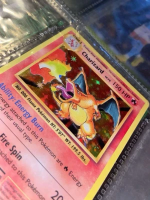Charizard Pokémonkort holografiskt - Säljer ett holografiskt Charizard Pokémonkort med gul ram och färgstark illustration. Kortet har 150 HP och visar Charizard i en eldig pose. Texten är på engelska och kortet har detaljer om attacker och förmågor. Perfekt för samlare eller Pokémonfans.