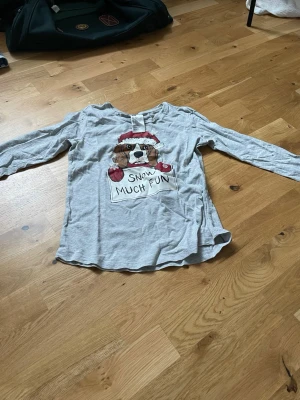 Grå långärmad topp med hundmotiv - Grå långärmad topp från H&M med ett gulligt hundmotiv i tomteluva och röda vantar samt texten 'Snow Much Fun' framtill. Perfekt för vintersäsongen och jul med sitt lekfulla tryck. Tillverkad i mjuk bomull för skön känsla.