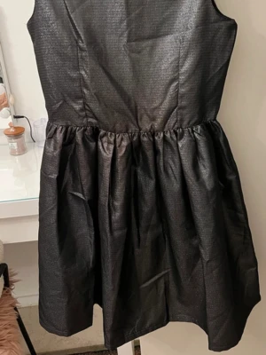 Svart glittrig kortklänning - Säljer en svart glittrig kortklänning med ärmlös design och rund hals. Klänningen har en markerad midja och ett utställt kjolparti som ger en snygg siluett. Dragkedja bak för enkel påklädning. Perfekt för dig som vill sticka ut lite extra!