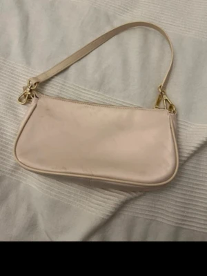 Ljusbeige axelremsväska i satin - Trendig ljusbeige handväska i satinliknande material med avtagbar axelrem och guldfärgade metalldetaljer. Väskan har en enkel, avlång form och stängs med dragkedja upptill. Perfekt för dig som gillar minimalistisk och stilren design.