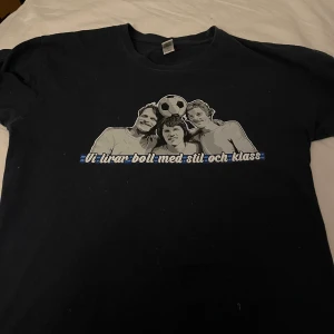 Sofialäktaren tisha - Svart t-shirt med ett stort tryck på bröstet som föreställer två personer och en fotboll. Under motivet står texten 'Vi lirar boll med stil och klass' i vitt och blått. Klassisk rund hals och korta ärmar. Perfekt för dig som gillar fotboll och streetwear.