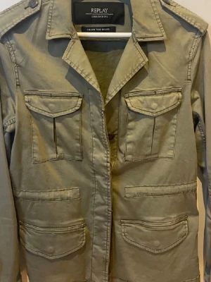 Olivgrön overshirt från Replay - Stilren olivgrön overshirt från Replay med fyra fickor framtill och axelklaffar. Klassisk krage och knappstängning. Tillverkad i slitstark bomull, perfekt för lager på lager. Svart Replay-märke på ärmen ger en snygg detalj.