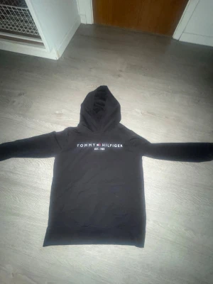 Svart hoodie från Tommy Hilfiger - Snygg svart hoodie från Tommy Hilfiger med klassisk logga och texten 'EST. 1985' på bröstet. Hoodien har huva och långa ärmar, samt en mjuk insida. Detaljer i rött, vitt och blått på insidan av huvan ger en extra touch. Perfekt för en avslappnad stil.