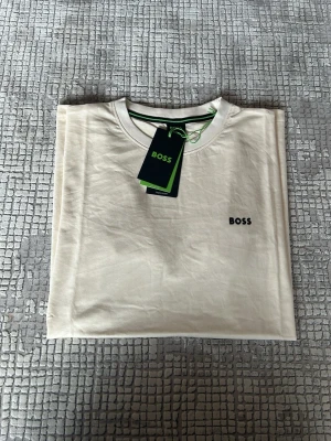 Vit kortärmad t-shirt från BOSS - Snygg vit t-shirt från BOSS med diskret logga broderad på bröstet. Klassisk rund halsringning och korta ärmar. Tillverkad i mjuk bomull för en skön känsla. Perfekt basplagg till garderoben.