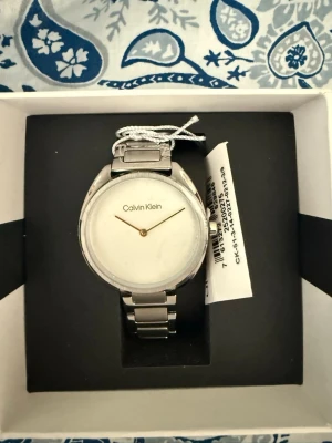 Silverfärgad klocka från Calvin Klein - Stilren klocka från Calvin Klein med rund, minimalistisk urtavla i vitt och silverfärgat armband i metall. Klassisk design utan siffror och med tunna visare. Perfekt accessoar för dig som gillar enkel och modern stil.