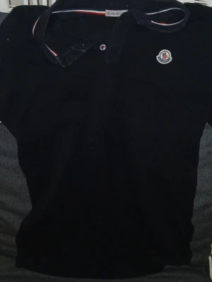 Svart Moncler pikétröja - Klassisk svart pikétröja från Moncler med korta ärmar och knappslå framtill. Tröjan har Monclers ikoniska logga på bröstet och detaljer i rött, vitt och blått på kragen. Tillverkad i mjuk bomull för en bekväm känsla. Den är lite smutisgt bara men annara helt ny inga hål eller något