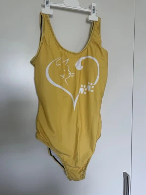 Gul baddräkt med kattmotiv - Snygg gul baddräkt med vitt tryck av en katt i hjärtform och tassavtryck på framsidan. Klassisk modell med breda axelband och rundad halsringning. Perfekt för dig som älskar djur och vill sticka ut på stranden.Helt ny med etikett 