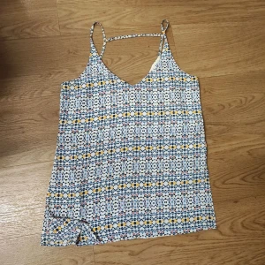 Mönstrad linnetopp med smala band - Säljer en tunn och luftig linnetopp med smala axelband och v-ringning. Toppen har ett färgglatt geometriskt mönster i vitt, blått och gult.