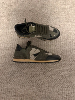 Valentino Rockrunners (Helt nya) - Snygga sneakers från Valentino i militärgrönt camomönster med detaljer i mocka och svart läder. Skorna har beige sula och klassisk låg siluett. Perfekta för dig som vill sticka ut med stilrena och trendiga detaljer.