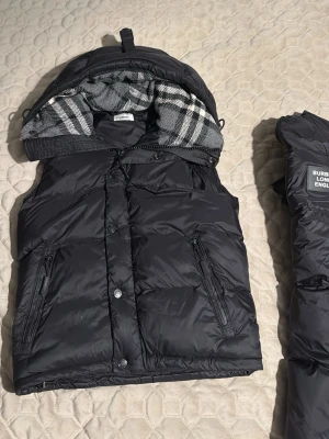 Svart dunväst från Burberry - Snygg svart dunväst från Burberry med avtagbara armar så man kan ha den som en väst och även en jacka och klassiskt rutigt foder. Västen har två dragkedjefickor framtill och tryckknappar. Materialet är glansigt och quiltat för extra värme. Perfekt för lager på lager och streetwear-stil. Den har ett litet hål som jag visa på sista bilden man ser inte det när man knäpper knapparna
