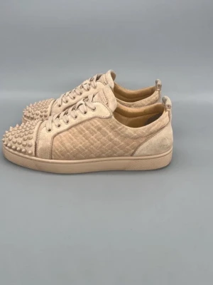 Beige sneakers med nitar från Louboutin - Säljer ett par beige sneakers från Christian Louboutin med coola nitar på tån och quiltat mönster på sidorna. Skorna har snörning, låg profil och är tillverkade i mocka för en lyxig känsla. Perfekta för dig som vill sticka ut med din stil.