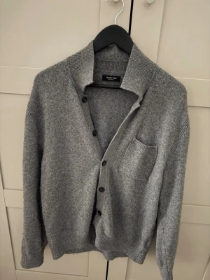 Grå kofta från Massimo dutti - Mysig grå kofta från Holzweiler med knappar framtill och en bröstficka. Koftan har ribbad krage, muddar och är stickad i ett mjukt material. Perfekt för lager på lager och enkel att matcha med olika outfits. ‼️Hör av er så kan vi snacka om priset!!