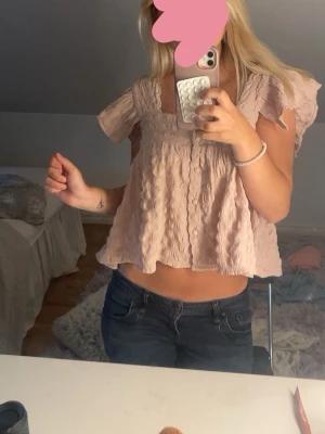 Ljusrosa volangblus med kort ärm - Supersöt ljusrosa blus med volangdetaljer och korta ärmar. Blusen har en luftig, loose passform och är tillverkad i ett tunt, strukturerat tyg. Perfekt för varma dagar och ger en romantisk vibe till din outfit.