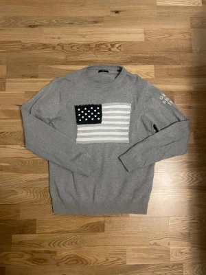 Grå tröja med amerikansk flagga Gant - Snygg grå tröja från Gant med amerikansk flagga i vitt och svart på bröstet. Rund halsringning och ribbade muddar. Diskret Gant-text på ena ärmen. Perfekt för dig som gillar klassisk och stilren design.