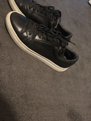 Svarta  sneakers i läder - Stilrena svarta sneakers i läder med vita sulor och svarta skosnören. Klassisk modell som passar till det mesta och har en enkel, clean design. Perfekta för dig som gillar minimalistisk stil. Har inte kartongen till skorna.