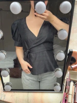 Svart omlottopp med puffärm - Snygg svart omlottopp med puffärmar och knyt i midjan. Toppen har v-ringning och korta ärmar som ger en trendig siluett. Perfekt att styla med jeans eller kjol för en cool look. Armén har förlängts med en bit tyg för att den var för liten, sista bilden visar