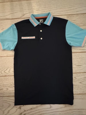 Blå pikétröja från FootJoy - Snygg pikétröja från FootJoy i mörkblått med ljusblå ärmar och krage. Tröjan har vita och röda detaljer på krage, ärmslut och bröstficka. Klassisk pikéknäppning med tre knappar och en liten bröstficka med randig kant.