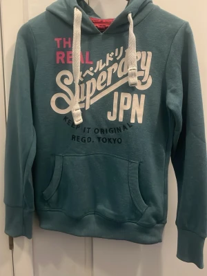 Blå Superdry hoodie med tryck - Fin blå hoodie från Superdry med stort vitt och rosa tryck på bröstet och japanska tecken. De. Är i storlek S men är lite stor i storleken. Som man ser på bilden har materialet blivit lite luddigt. Lite svårt att förklara färgen men den är blå/grå. Kan diskutera pris 🤗