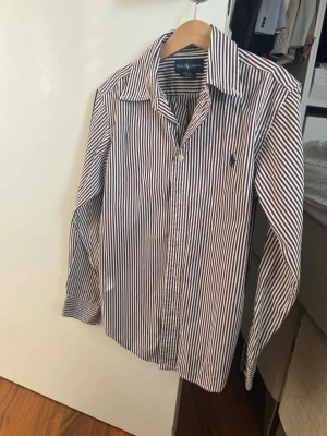 Randig skjorta från Ralph Lauren - Klassisk skjorta från Ralph Lauren med smala vertikala ränder i blått och vitt. Skjortan har lång ärm, krage och knäppning framtill samt den ikoniska broderade loggan på bröstet. Tillverkad i mjuk bomull för en bekväm känsla.