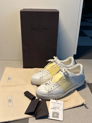 Valentino Garavani vita sneakers - Snygga vita sneakers från Valentino Garavani med breda ljusgula band över ovandelen och klassisk låg siluett. Skorna har vita snören, silverfärgade nitar och är tillverkade i slätt läder. Kommer med originalkartong och dustbags. Den ända lilla deffekten är en röd fläck men den syns knappt