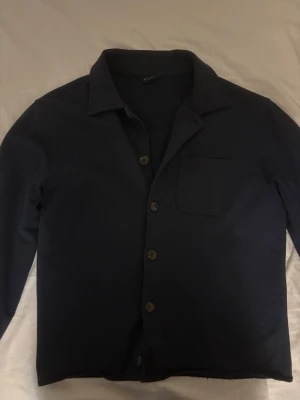 Mörkblå Overshirt från Riley - Stilren mörkblå overshirt från Riley med klassisk krage, knappstängning framtill och en bröstficka. Overshirten har raka ärmar och är tillverkad i ett mjukt, bomullsliknande material som ger en avslappnad look.