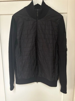 Svart cardiganjacka från J.Lindeberg - Stilren svart cardiganjacka från J.Lindeberg med quiltad front och stickade ärmar. Jackan har hög krage, hel dragkedja och ribbade muddar. Perfekt och trendig vårjacka! Mycket fint skick. Passar Medium-Large.