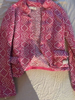 Rosa mönstrad cardiganjacka - Säljer en rosa cardiganjacka med vitt geometriskt mönster. Jackan har öppen front, två fickor och en mjuk, stickad känsla. Perfekt för dig som vill ha något färgglatt och unikt i garderoben.