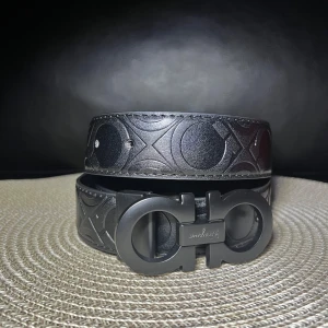 Bälte från Salvatore Ferragamo 105 cm - Snyggt svart skärp från Salvatore Ferragamo med präglat mönster och ikoniskt dubbel-Gancio-spänne i matt svart metall. Skärpet har en bred design och är tillverkat i skinn, perfekt för att lyfta vilken outfit som helst.