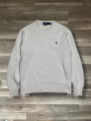 Vit stickad tröja från Polo Ralph Lauren - Klassisk vit stickad tröja från Polo Ralph Lauren med rund halsringning och ribbade muddar. Tröjan har den ikoniska blå broderade loggan på bröstet och är tillverkad i ett mjukt, tjockt material. Perfekt för lager på lager under kyliga dagar.