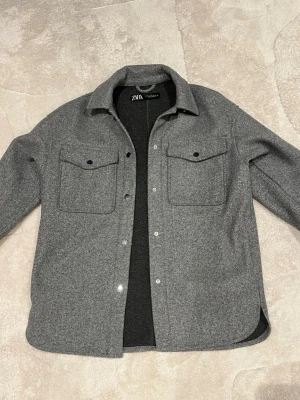 Grå overshirt från ZARA i ullmix - Stilren grå overshirt från ZARA med klassisk krage och två stora bröstfickor med knappar. Jackan har tryckknappar framtill och är tillverkad i ett mjukt ullblandat material. Perfekt lager-på-lager-plagg för en avslappnad look. Den har lite noppror på vissa ställen, men inget som direkt syns! 