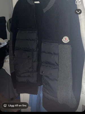 Svart cardiganjacka från Moncler - Snygg svart cardiganjacka från Moncler med stickade ärmar och vadderade paneler framtill. Jackan har två stora fickor med knappar och dragkedja framtill. Moncler-logga på ärmen och ribbade muddar. Perfekt för lager-på-lager och streetstyle.