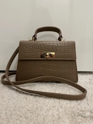 Brun croco handväska med axelrem - Snygg brun handväska i croco-mönstrat konstläder med guldfärgade detaljer och vridlås. Väskan har både ett topphandtag och en lång axelrem så du kan bära den på flera sätt. Perfekt storlek för det viktigaste och ger en trendig touch till din outfit.