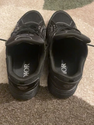 Svarta Dior sneakers med vita sömmar - Säljer ett par svarta D•or sneakers med tydliga vita kontrastsömmar och klassisk snörning. Skorna har en chunky sula och meshdetaljer för extra komfort. D•or-loggan syns på sidan och innersulan. Perfekta för dig som vill ha en stilren och exklusiv look.