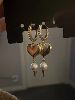 Guldörhängen med hjärta och pärla - Snygga örhängen i guldfärg med hjärtformade hängen och vita pärlor. Örhängena har små gnistrande stenar på ringen och en cool spetsig detalj under pärlan. Perfekta för att lyfta vilken outfit som helst.