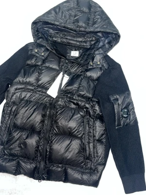 Svart dunväst med huva C.P. Company - Svart dunväst från C.P. Company med glansigt quiltat material, stickade svarta ärmar och huva. Västen har dragkedja framtill, två sidofickor och den ikoniska linsen på ärmen. Perfekt för dig som gillar streetwear och vill ha en modern look.